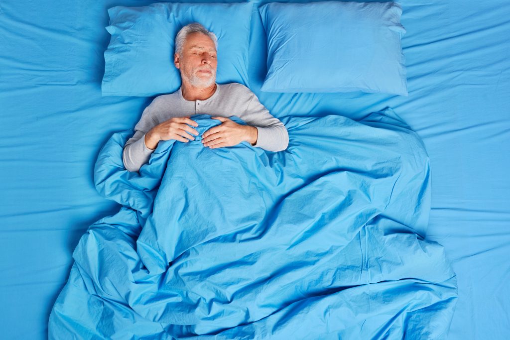 High angle view of calm bearded grey haired senior man sleeps peacefully in bed enjoys pleasant dreams feels tired after hard day lives alone poses on soft blue pillow. Early morning concept